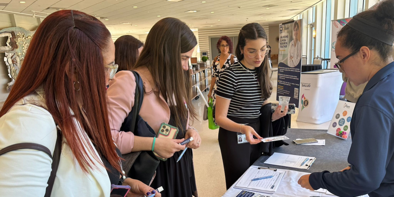 ISCORE in the SD2 A group of people gathered around an informational booth at a conference or event. The table is covered with brochures, flyers, and pens. One person is holding a flyer, another is using a smartphone. Posters and banners in the background provide information about clinical research and health services. The event is indoors with large windows letting in natural light.