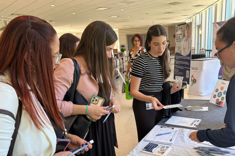ISCORE in the SD2 A group of people gathered around an informational booth at a conference or event. The table is covered with brochures, flyers, and pens. One person is holding a flyer, another is using a smartphone. Posters and banners in the background provide information about clinical research and health services. The event is indoors with large windows letting in natural light.