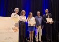 A group of five individuals stands on a stage during an award ceremony. Two recipients in the center hold certificates and small gift boxes. A sign in the foreground reads: “The Alliance – 4th Scientific Day: Advances in Health through Implementation Science: Research in Real-World Practice and Community Settings, May 7, 2025, Caribe Hilton Hotel.”