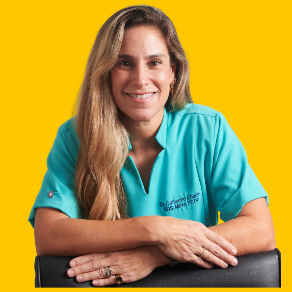 Dr. Catherine Chacín, wearing a teal shirt embroidered with her name and credentials (DDS, MPH, FETP), smiles confidently with her arms resting on a surface, against a bright yellow background.
