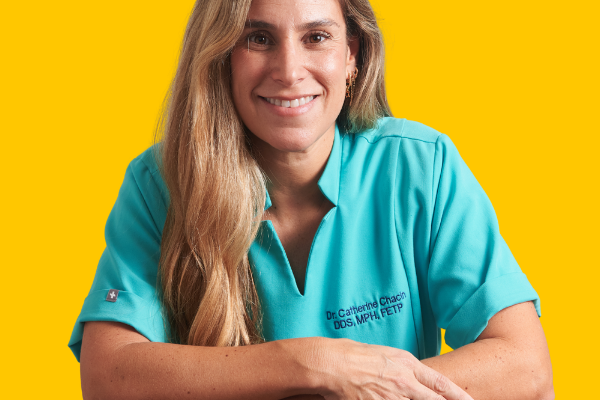 Dr. Catherine Chacín, wearing a teal shirt embroidered with her name and credentials (DDS, MPH, FETP), smiles confidently with her arms resting on a surface, against a bright yellow background.